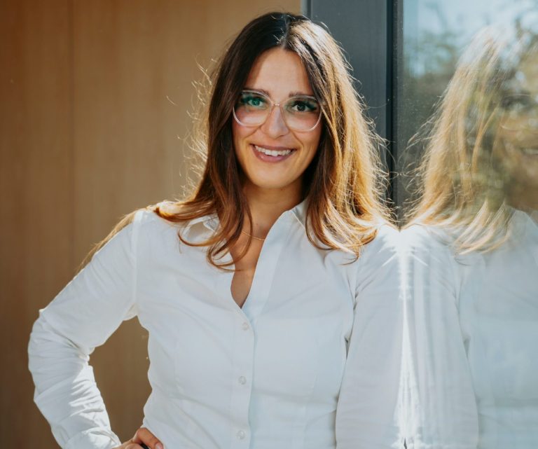 Luisa Wichter Frau mit langen Haaren in weißem Hemd lächelt, steht vor einem Fenster.